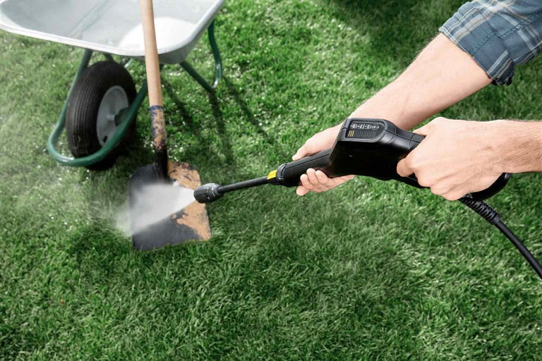 Osoba čistí lopatu vysokotlakým čističem na zahradě, s kolečkem v pozadí.