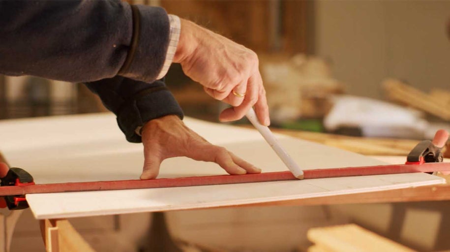 Osoba označuje čáru tužkou a pravítkem na dřevěné desce upevněné svorkami.