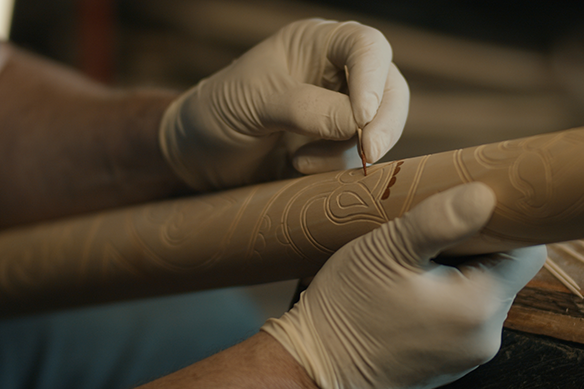 Detailní záběr rukou v ochranných rukavicích, které opracovávají dřevěnou tyč nástrojem