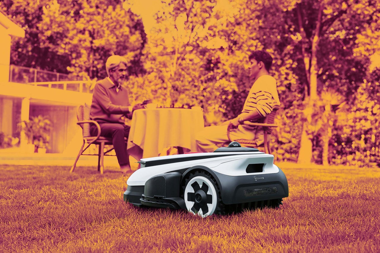 Robotická sekačka Ecovacs na trávníku v zahradě, v pozadí sedí dvě osoby u stolu.