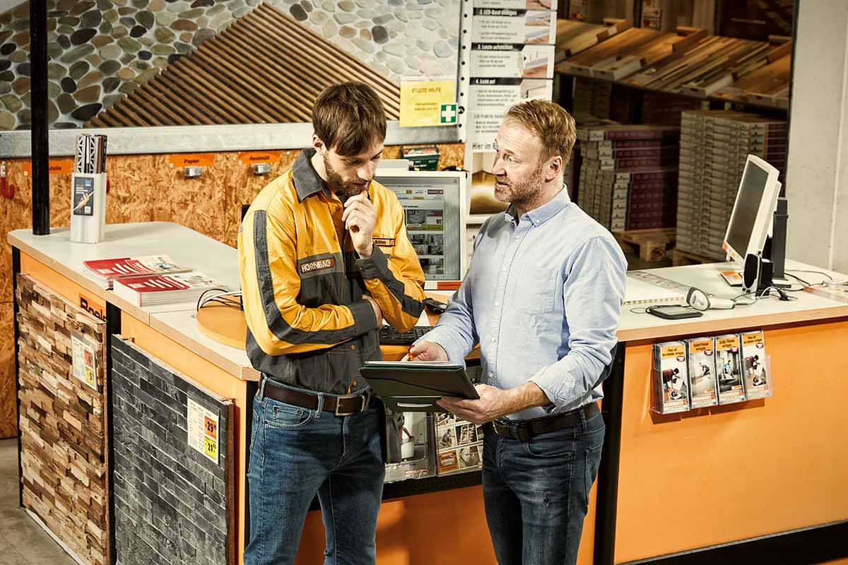 Zaměstnanec Hornbachu v uniformě radí zákazníkovi u servisního pultu v hobbymarketu při prohlížení tabletu.