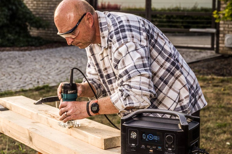 Muž používá horní frézku na dřevěné desce venku, s přenosnou napájecí stanicí v popředí.