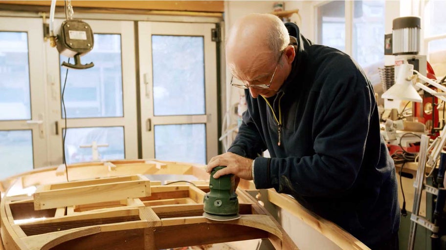 Muž brousí dřevěnou loď elektrickou bruskou v dílně.
