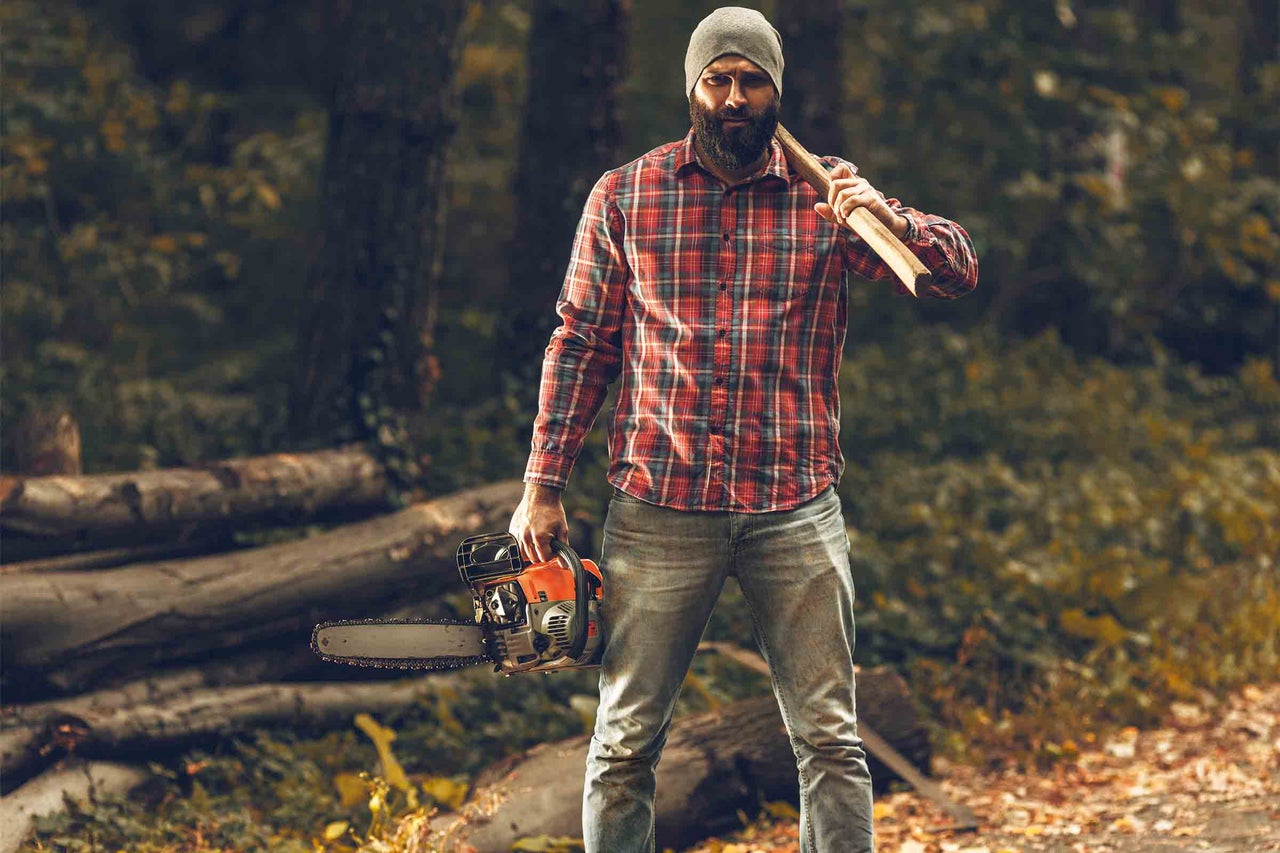 Muž se sekerou na rameni a motorovou pilou v ruce v lese