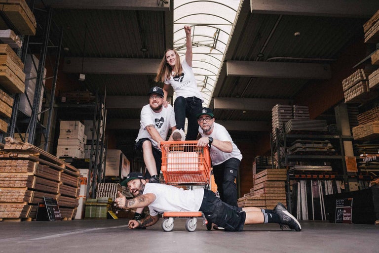 Čtyři lidé pózují s nákupním vozíkem ve skladu se stavebními materiály. Jeden muž leží na skateboardu.