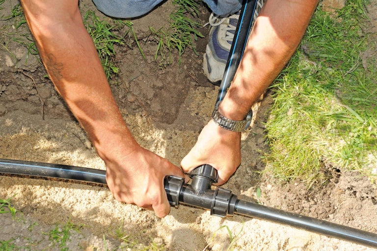 Osoba spojuje černou plastovou trubku s T-kusem pro zavlažování zahrady.