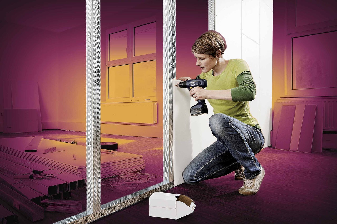 Žena připevňuje sádrokartonovou desku akumulátorovým šroubovákem na kovový rám v interiéru.