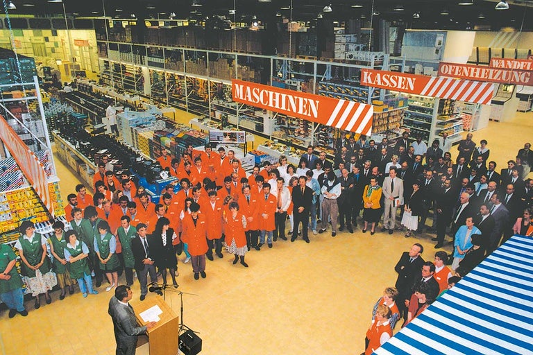 Velká skupina lidí stojí v obchodě Hornbach pod cedulemi pro pokladny, stroje a kamna.