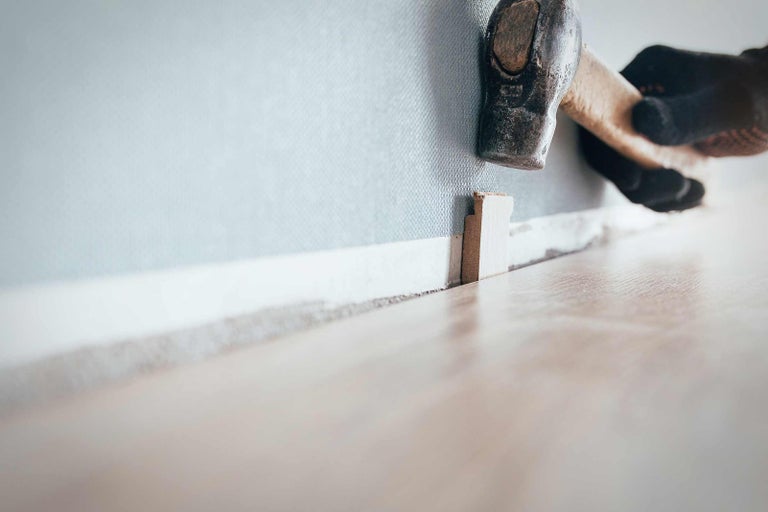 Řemeslník připevňuje kladivem soklovou lištu ke stěně.