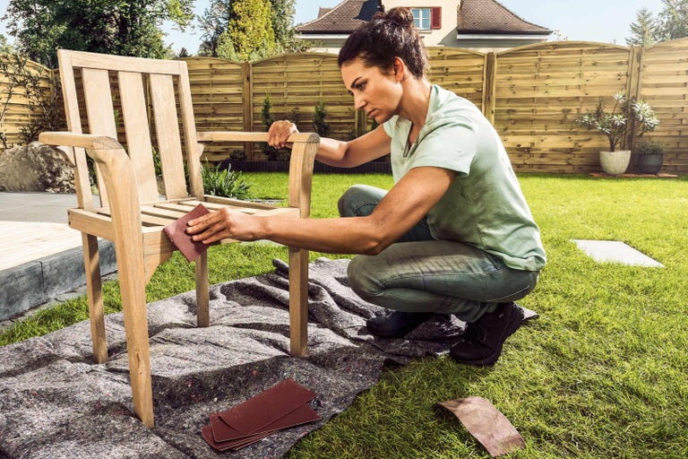 Žena brousí dřevěnou židli v zahradě brusným papírem