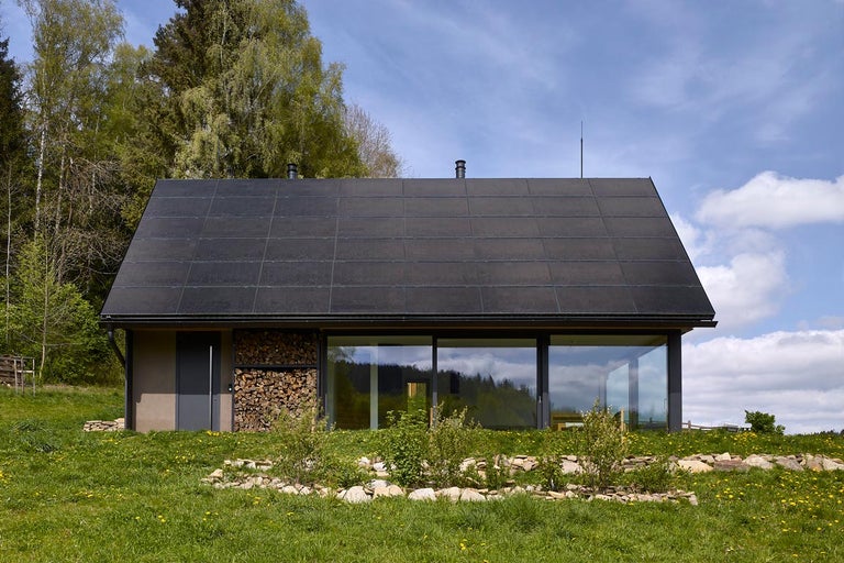 Moderní dům se solárními panely na střeše, skladem dřeva a velkými okny.