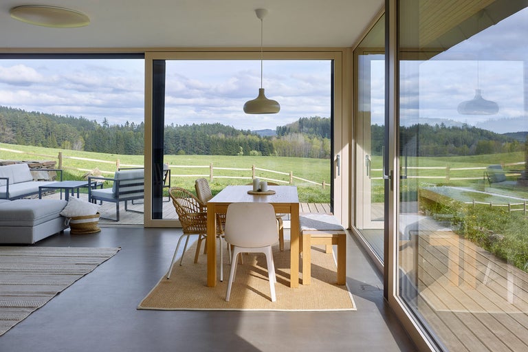 Světlý obývací pokoj s jídelním stolem, židlemi, lavicí a velkým oknem s výhledem do přírody.