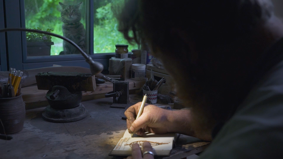 Muž kreslí tužkou do zápisníku na stole s nářadím, zatímco se z okna dívá kočka.