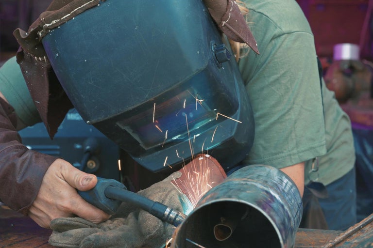 Osoba svařuje kovovou trubku s ochrannou maskou a rukavicemi.