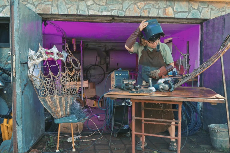 Dílna s kovovou sochařkou, která má na sobě svářečskou kuklu a pracuje na kovové soše.