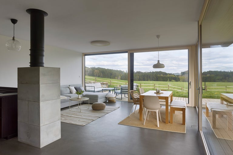 Světlý obývací pokoj s krbem, pohovkou a jídelním stolem před velkým oknem s výhledem do přírody.