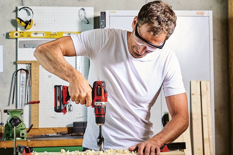 Muž vrtá aku vrtačkou s nástavcem otvor do kusu dřeva. Má ochranné brýle.