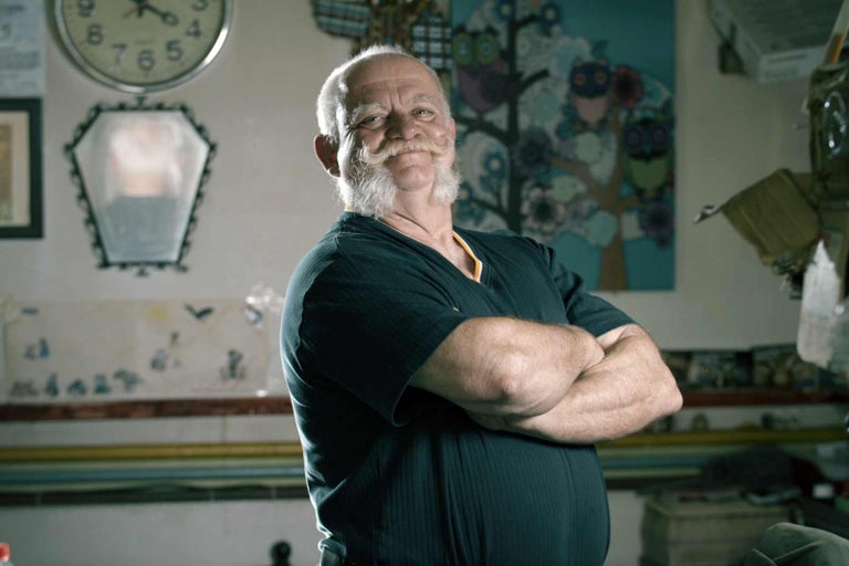 Portrét muže s vousy a zkříženýma rukama v místnosti s hodinami, zrcadlem a obrazem.