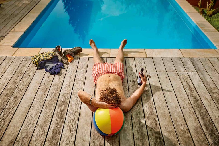 Muž relaxuje u bazénu na dřevěné terase s pitím a míčem