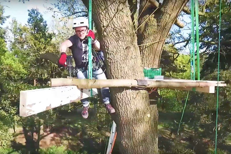 Muž s ochrannou přilbou pracuje s vrtačkou na domku na stromě