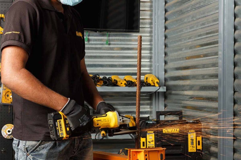 Muž řeže úhlovou bruskou kus kovu, přičemž létají jiskry. V pozadí je nářadí a logo DeWalt.