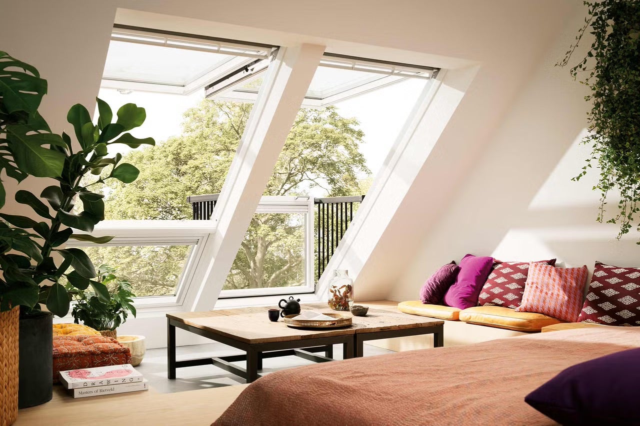 Světlý podkrovní pokoj se dvěma otevřenými střešními okny tvořícími balkon, nízkým dřevěným stolem a pokojovými rostlinami.
