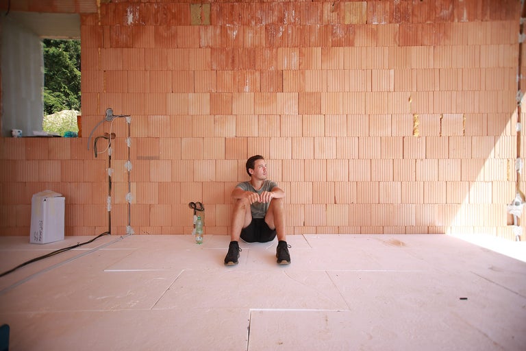Interiérový snímek hrubé stavby s cihlovými stěnami a izolovanou podlahou, na které sedí muž