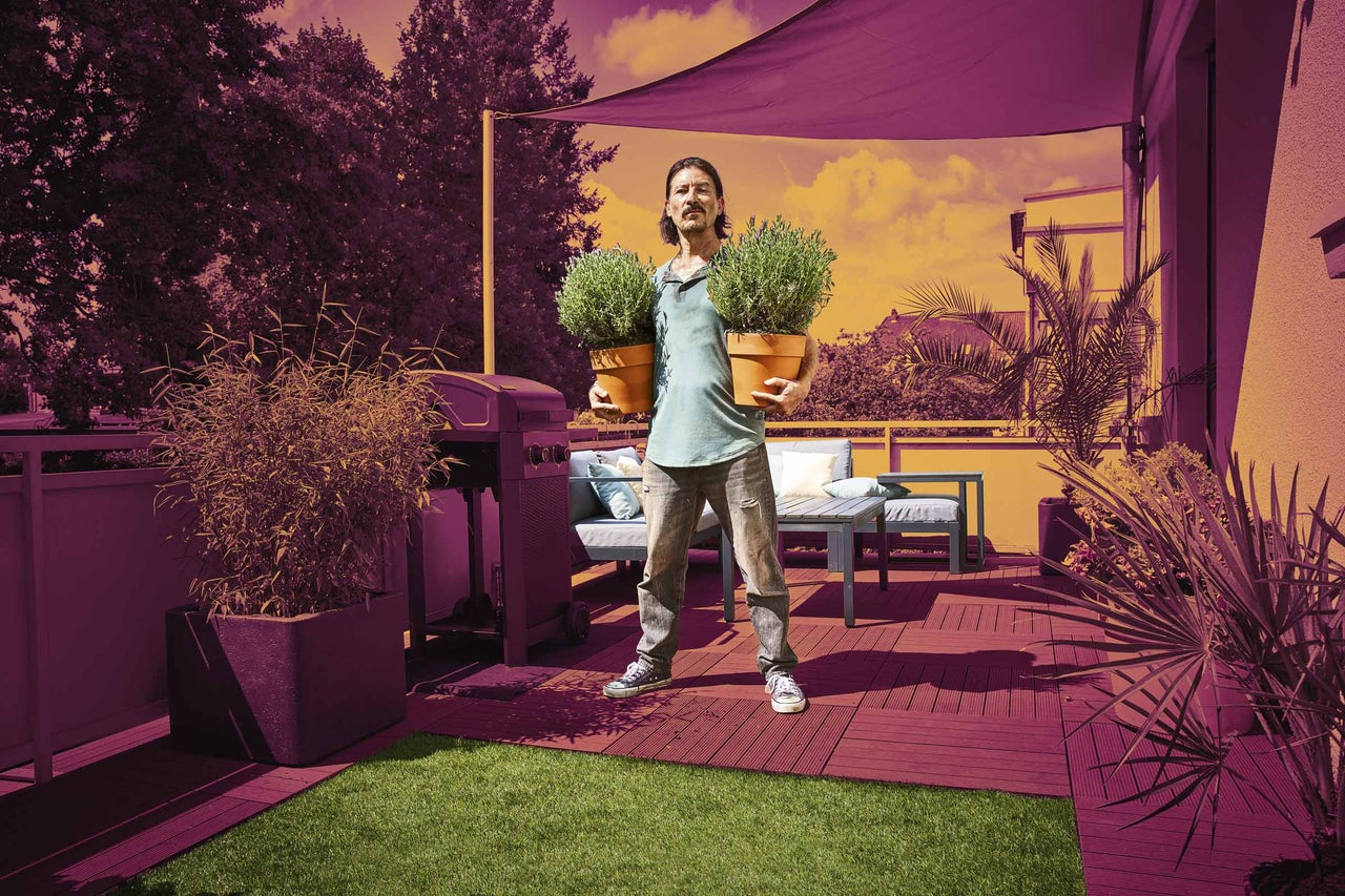 Muž s levandulí na terase s dřevěnou dlažbou, umělým trávníkem, zahradním nábytkem, plynovým grilem a stínicí plachtou.