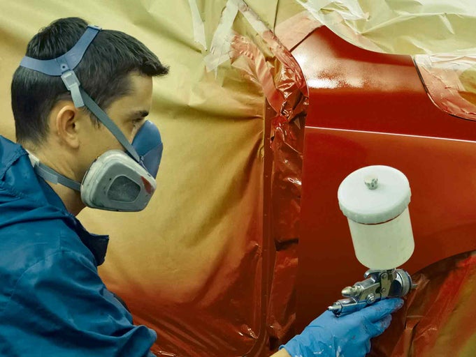 Řemeslník lakuje auto pomocí stříkací pistole. Má na sobě ochrannou masku a rukavice.