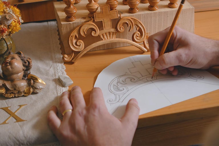 Osoba kreslí ozdobu tužkou na papír, vedle vyřezávané dřevěné ozdoby a figurky anděla.