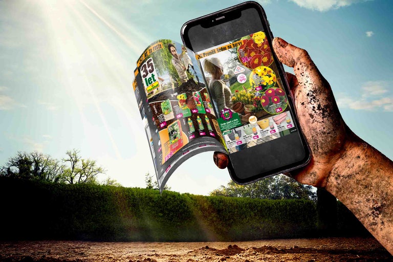 Špinavá ruka drží smartphone s reklamou Hornbach se zahradními potřebami, v pozadí zahrada a obloha