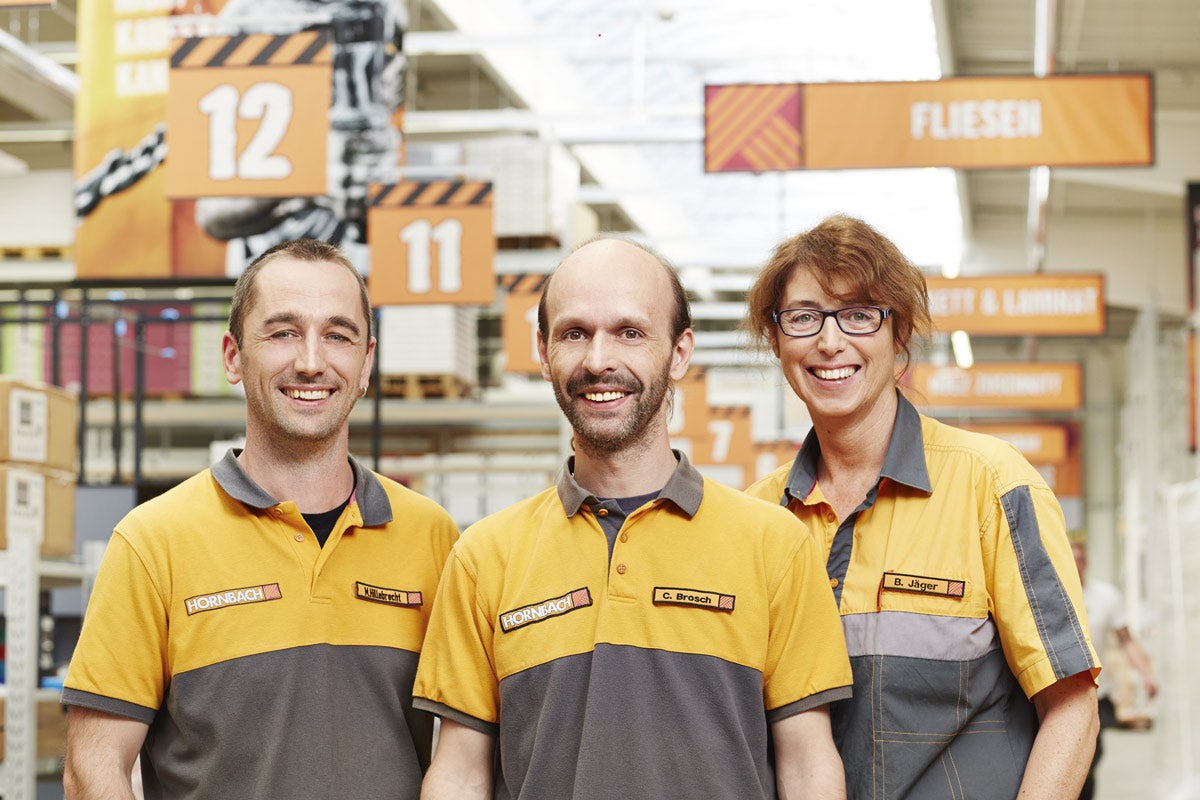 Tři zaměstnanci Hornbach v pracovním oblečení před regály se stavebním materiálem.