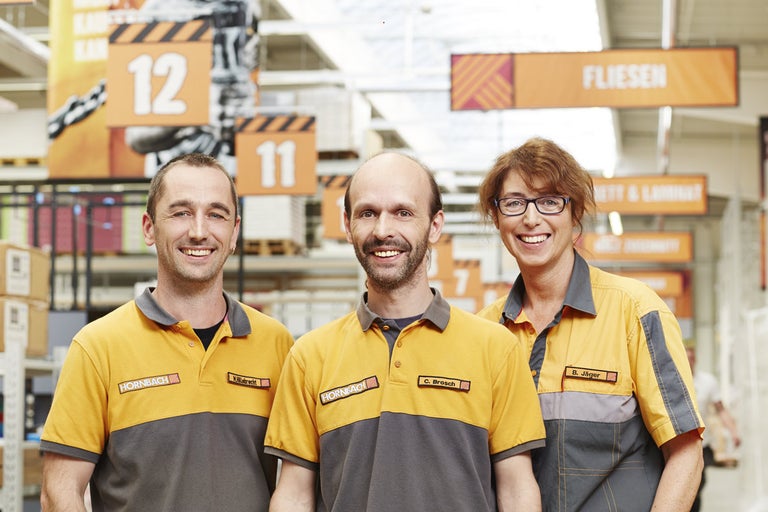 Tři zaměstnanci Hornbach v pracovním oblečení před regály se stavebním materiálem.