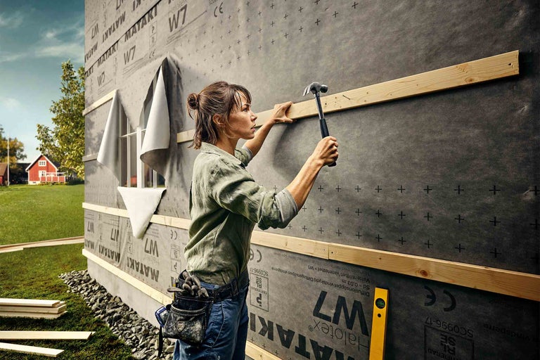 Žena přibíjí dřevěnou lištu na stěnu domu s větrnou membránou.