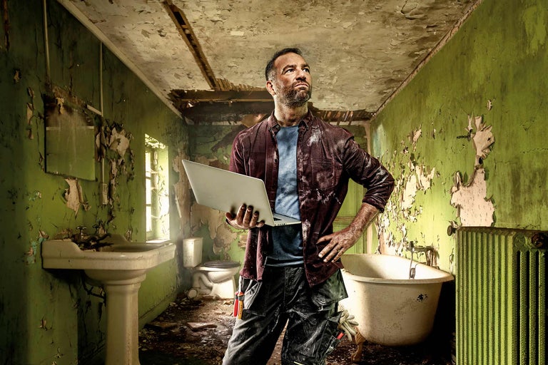 Řemeslník s notebookem v koupelně vyžadující rekonstrukci s vanou, toaletou a umyvadlem