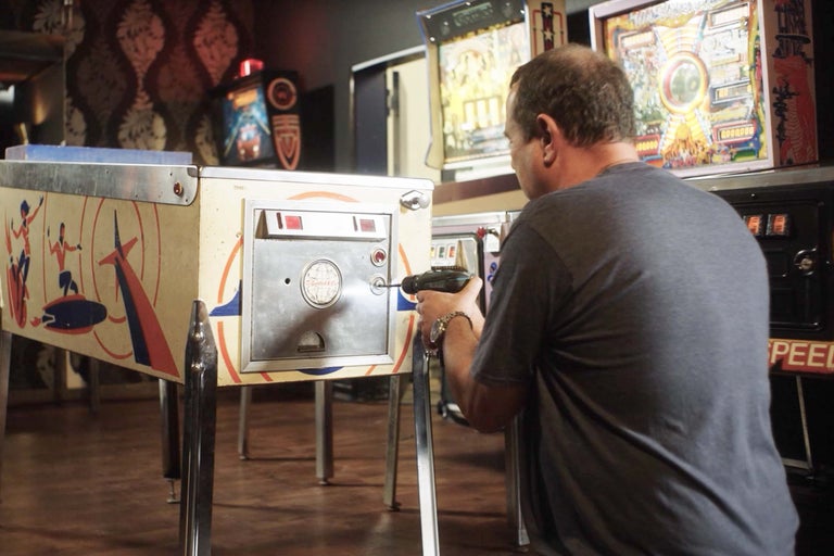 Muž opravuje pinballový automat pomocí akumulátorového šroubováku v herně