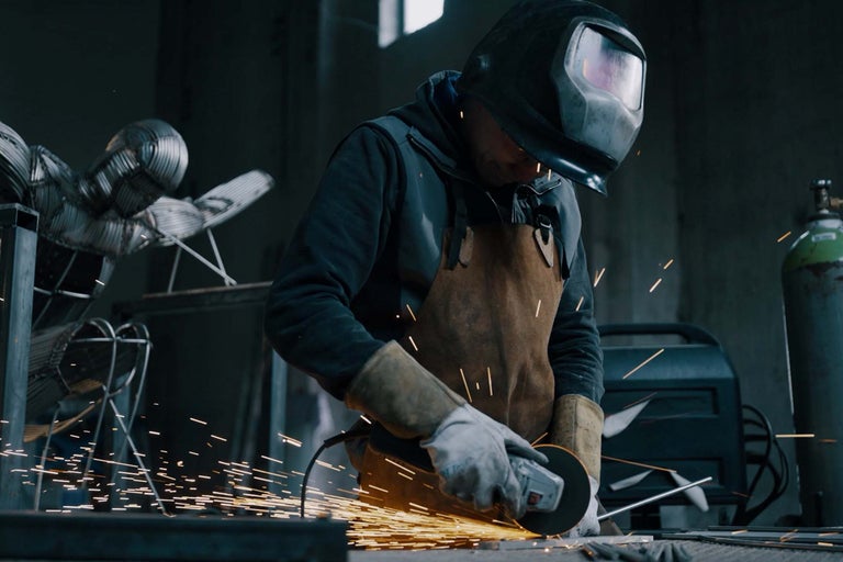 Muž s ochrannou helmou a zástěrou používá úhlovou brusku v dílně