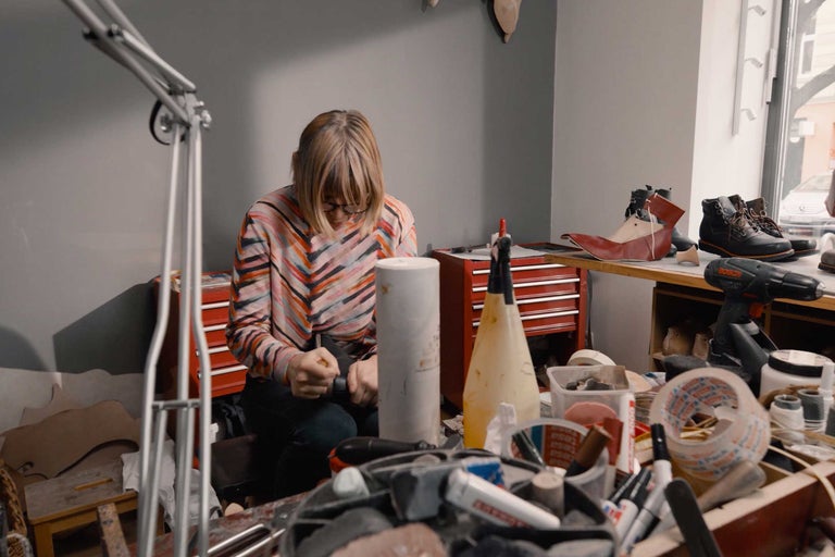 Žena pracuje ve své dílně na výrobě obuvi, na stole leží nářadí a materiál.