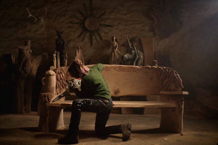 Žena klečí před dřevěnou lavicí a pracuje na ní s motorovou pilou. V pozadí jsou různé dřevěné sochy.