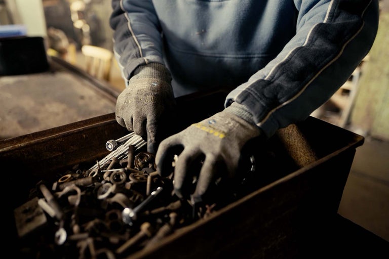 Osoba prohledává bednu se šrouby a maticemi v pracovních rukavicích.