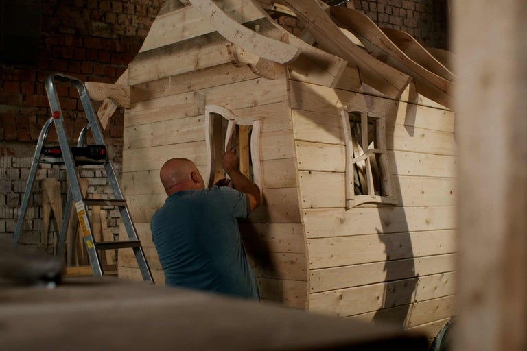 Dřevěný domeček pro děti staví muž. Vedle stojí žebřík.