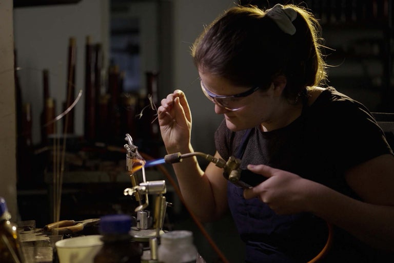 Žena s ochrannými brýlemi a pájecí lampou při zpracování skla.