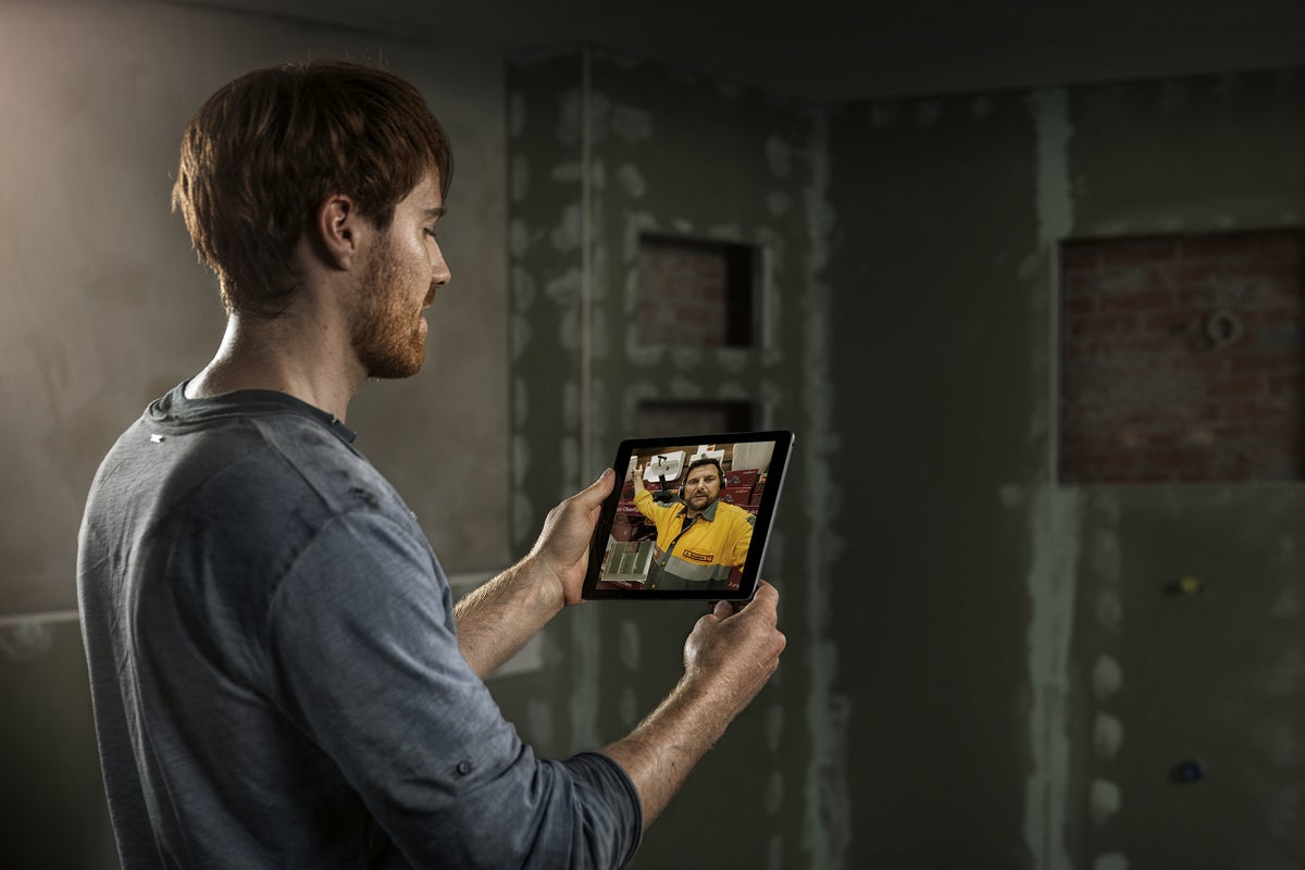 Muž drží tablet s videoporadenstvím před stěnou ve výstavbě.