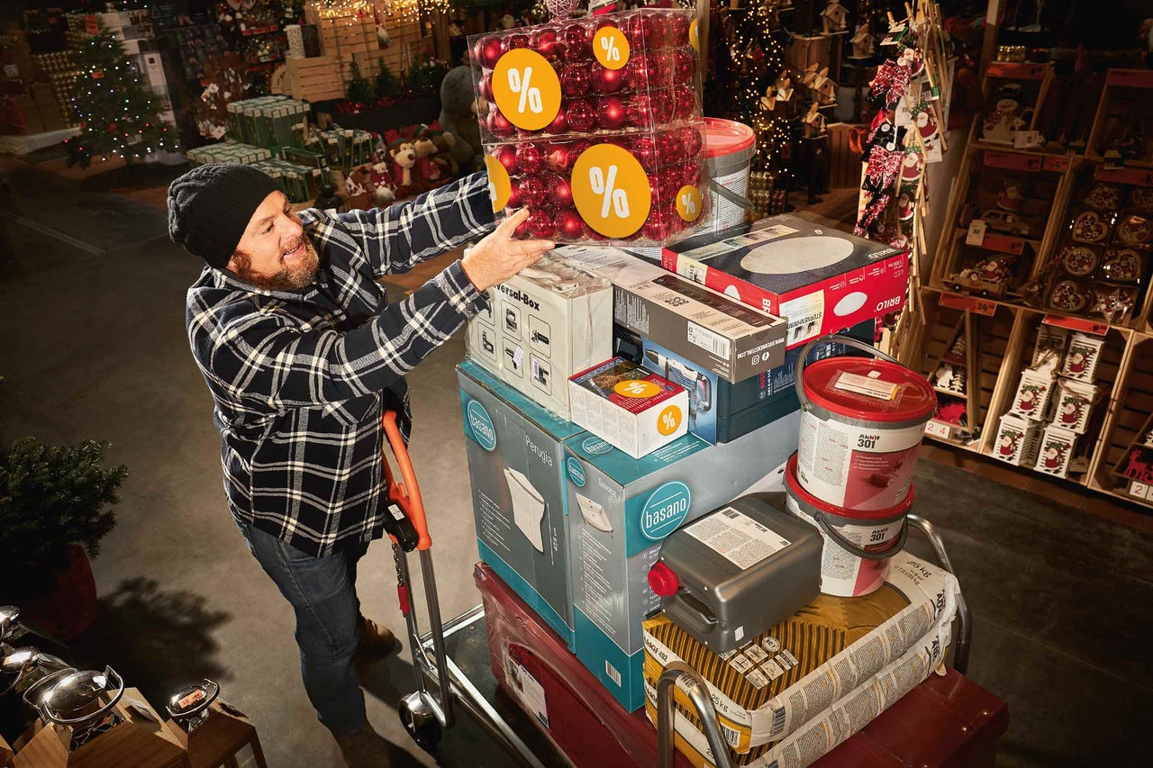 Muž v hobbymarketu Hornbach skládá nákupy, včetně vánočních koulí, barvy a stavebních materiálů, na vozík.