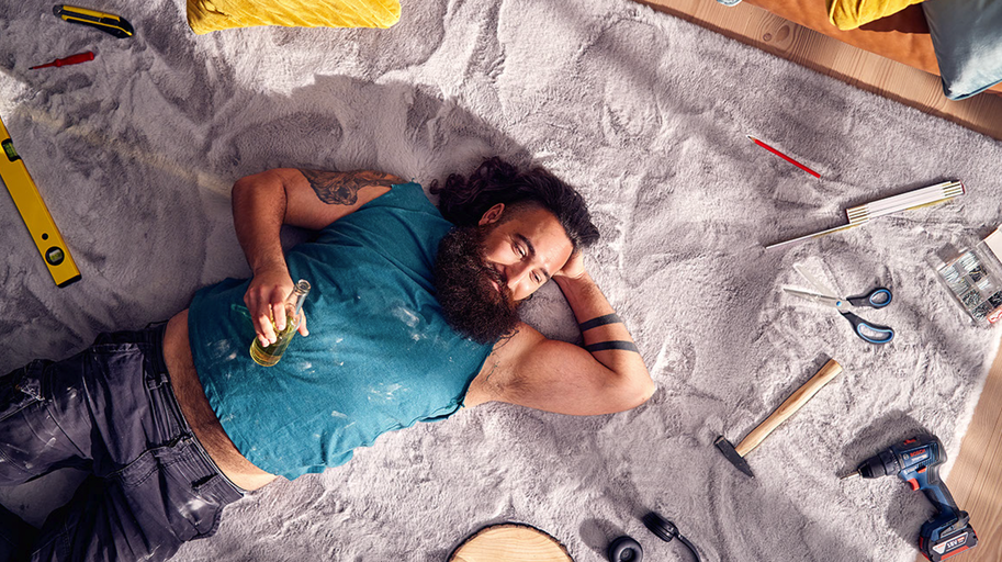 Muž leží vyčerpaný na koberci s nářadím a lahví v ruce