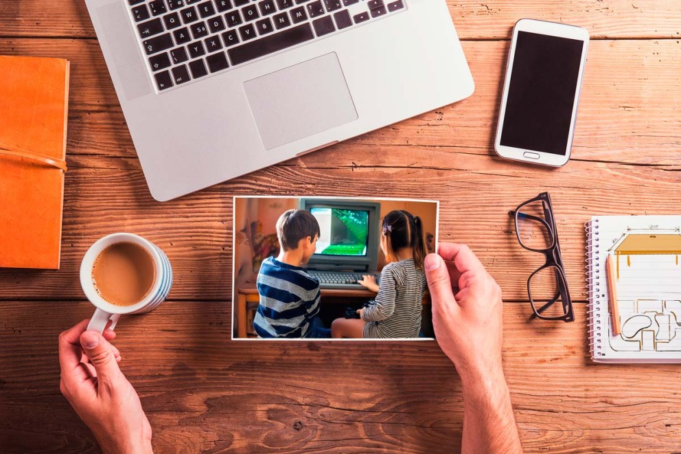 Pohled shora na stůl s notebookem, zápisníkem, brýlemi, chytrým telefonem, šálkem kávy a fotografií dvou dětí sedících před počítačem