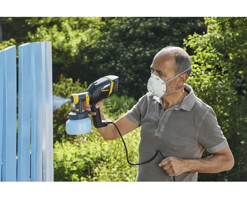 Osoba používá stříkací pistoli k natírání dřevěného plotu. Osoba má na sobě ochrannou masku.