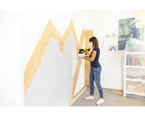 Žena stříká barvu na zeď pomocí stříkacího systému na stěnu, která je zakrytá maskovací páskou.