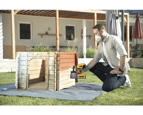 Muž natírá psí boudu na zahradě stříkací pistolí.