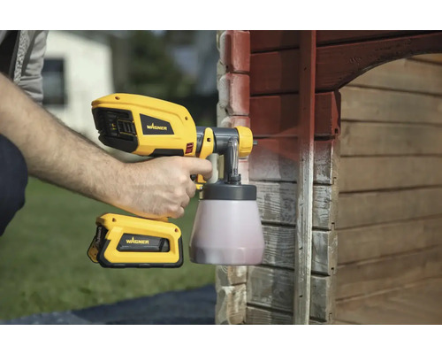 Muž používá stříkací pistoli Wagner k natírání dřevěného domu.